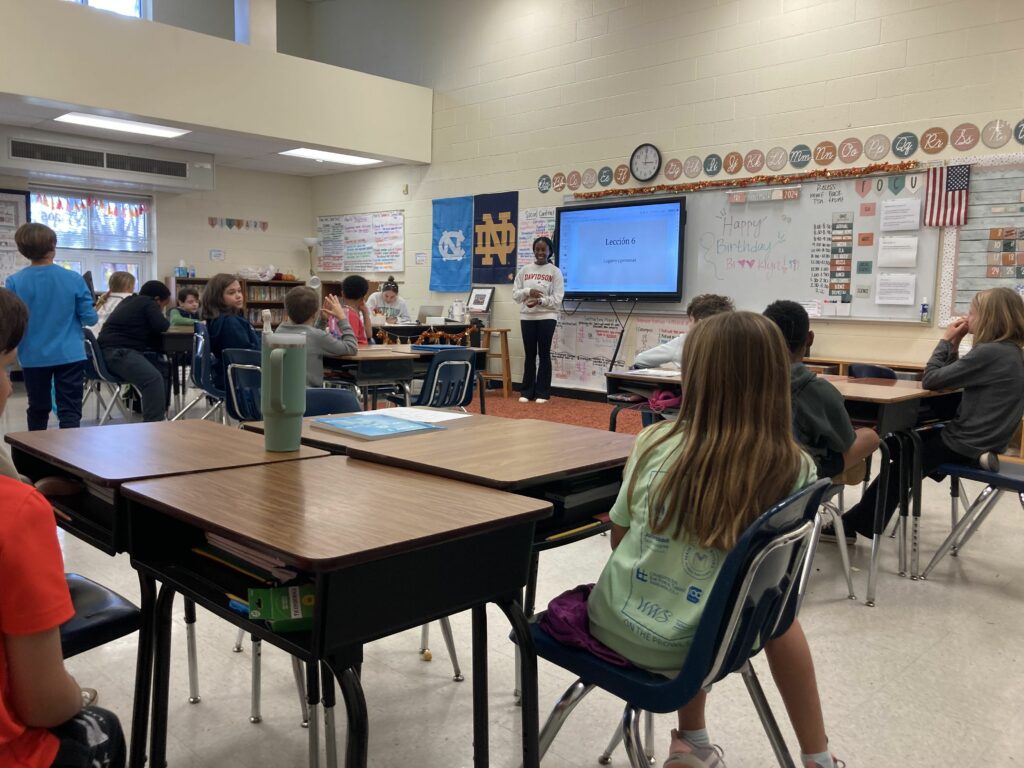 About 20 4th grade students sitting at their desks in small groups of four, facing the teacher (a 19 year old female in a davidson college sweatshirt smiling). There is a slide show of lección 6.